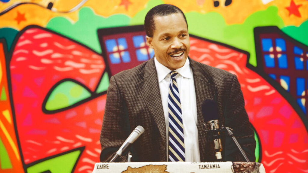 Joseph Holland at a podium with colorful art painted on the walls behind him.